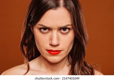 Face Portrait Of Young Angry Hateful Woman Looking At Camera At Studio Over Brown Background. Upset, Frustrated, Anger, Rage Female Facial Expression.