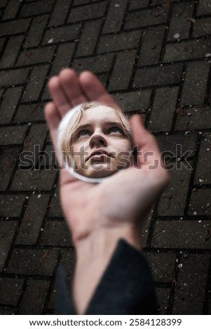 A face is reflected in a small handheld mirror, creating an artistic and surreal perspective against a paved background, symbolizing introspection, perception, and self-awareness