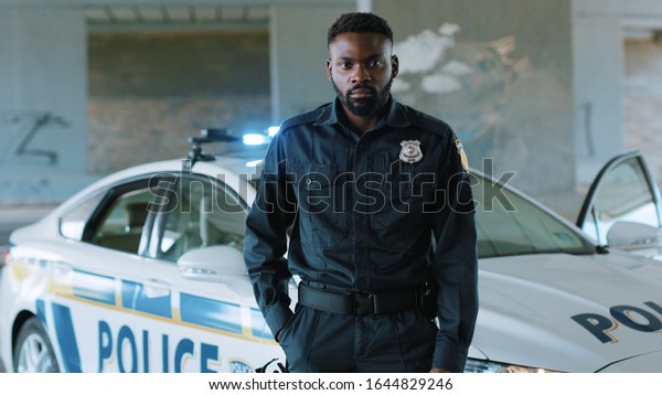 Face serious african american young man cops stand\
near patrol car look at camera enforcement happy officer police\
uniform auto safety security communication control policeman close\
up slow motion