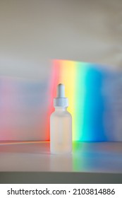 Face Serum In Matte Glass Bottle With A Dropper In Front Of Pastel Rainbow Prism Background. Front View