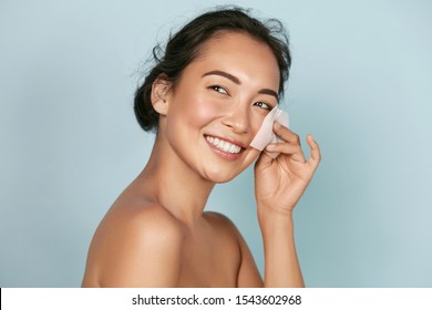 Face Skin Care. Smiling Woman Using Facial Oil Blotting Paper Portrait. Closeup Of Beautiful Happy Asian Girl Model With Natural Makeup Using Oil Absorbing Sheets, Beauty Product At Studio