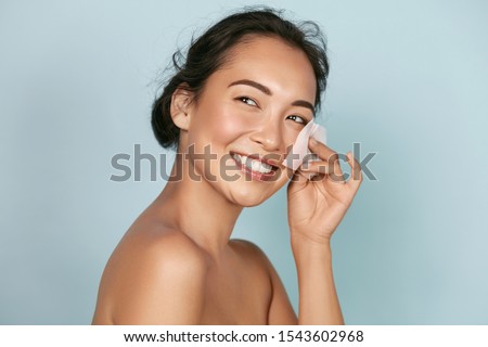 Face skin care. Smiling woman using facial oil blotting paper portrait. Closeup of beautiful happy asian girl model with natural makeup using oil absorbing sheets, beauty product at studio