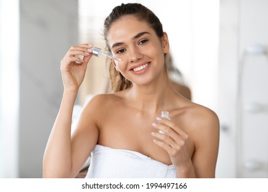 Face Skincare. Happy Woman Applying Facial Serum With Dropper Moisturizing Skin Smiling To Camera Standing Posing In Bathroom At Home. Domestic Skin Care Routine Concept. Beauty And Cosmetics