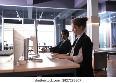 Face to face workstations in the bright office, both employees absorbed at their screens with warm spotlights and glass corridors receding behind them in a bright modern coworking office.