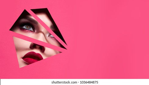 the face of a young beautiful girl with a bright make-up and with plump red lips peeks into a hole in pink paper.Fashion, beauty, make-up, cosmetics, beauty salon, style, personal care, geometry. - Shutterstock ID 1036484875