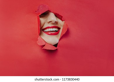 the face of a young beautiful girl with a bright make-up and with plump red lips peeks into a hole in pink paper.Lipstick,cosmetics,makeup, fashion, beauty,beauty salon.