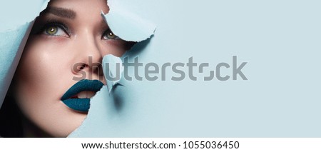 the face of a young beautiful girl with a bright make-up and puffy blue lips peers into a hole in blue paper.Fashion, beauty, make-up, cosmetics, hairstyle, beauty salon, boutique, discounts, sales. Foto d'archivio © 