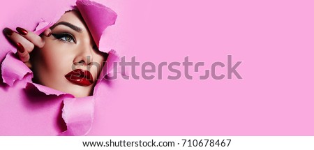the face of a young beautiful girl with a bright make-up and with plump red lips peeks into a hole in pink paper.Beauty, fashion, personal care,cosmetics, make-up,health, beauty salon, make-up artist.