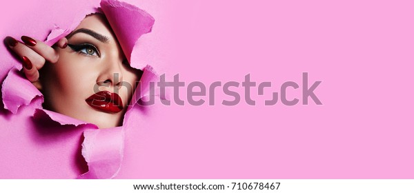 the face of a young beautiful girl with a bright\
make-up and with plump red lips peeks into a hole in pink\
paper.Beauty, fashion, personal care,cosmetics, make-up,health,\
beauty salon, make-up\
artist.