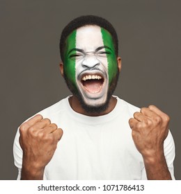 Face Of Young Screaming African-american Man Painted With Flag Of Nigeria. Football Or Soccer Team Fan, Sport Event, Faceart And Patriotism Concept. Studio Shot At Gray Background, Copy Space