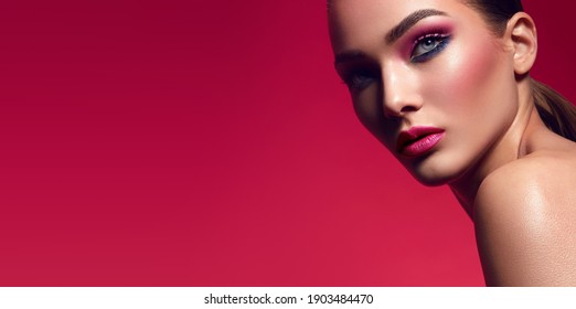 Face of a young woman with bright makeup on a pink background with gathered hair in a smooth ponytail.