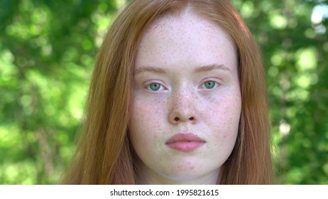 Face of a young woman with red hair and freckles on a background of green foliage. Natural beauty with freckles.