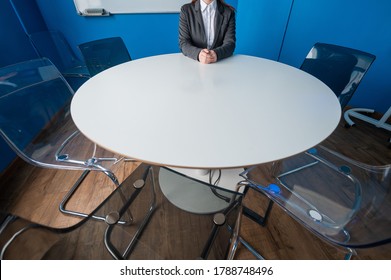 A faceless business woman in a suit sits alone at a round table in a meeting room. The female boss is waiting for her team in the boardroom.