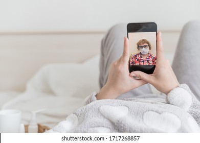 A faceless girl in quarantine lies in bed in her pajamas and makes a video call on the phone to her mother. Elderly woman in a medical mask on a smartphone screen. - Powered by Shutterstock