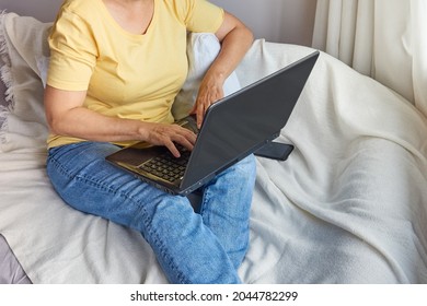 faceless image of an older woman in a yellow shirt uses a laptop while sitting on the couch, a pensioner working at a computer or studying at home, business, education online - Powered by Shutterstock