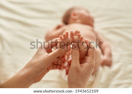 Faceless person making baby massage for infant lying on bed, mommy massaging new born baby with love, mum gently touching kid's feet for development of coordination of movements.