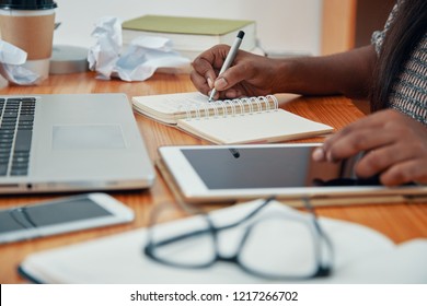 Faceless Shot Of Black Woman Taking Notes In Notepad While Using Tablet At Working Desk In Office