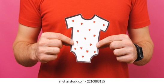 Faceless Shot Of A Male Father Holding Paper Onesie With Little Hearts Over Pink And Red Background.