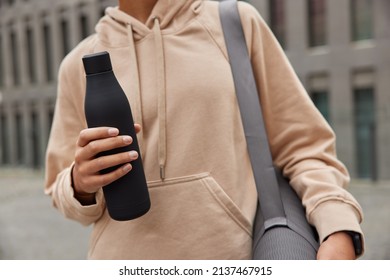 Faceless sporty athletic woman dressed in brown sweatshirt carries rolled rubber mat for workout holds sport bottle of water returns from gym after fitness training poses against blurred background.