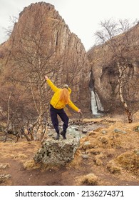 A Faceless Woman Stands In A Pose On A Stone Among Bare Birches Against The Backdrop Of Steep Cliffs And A Waterfall. Girl With Hair Instead Of A Face Jokey Photo