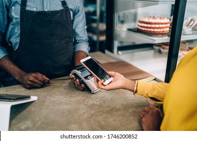 Faceless in yellow dress paying for order with mobile phone using payment terminal in cafe standing at counter with cakes and desserts 
