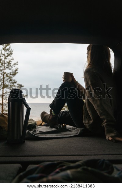 Faceless young girl in beige sweater enjoying\
seascape. Woman sitting in car trunk and drinking hot tea from\
thermos. Travelling by car. Freedom\
concept.
