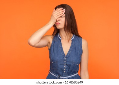 Facepalm. Distressed Regretful Brunette Woman In Denim Dress Covering Face With Hand, Feeling Shame And Sorrow About Defeat, Desperate Emotions. Indoor Studio Shot Isolated On Orange Background