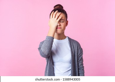 Facepalm, oh no! Portrait of unhappy teen girl with bun hairstyle in casual clothes standing with hand on head, feeling sorrow regret blaming herself for failure. studio shot isolated, pink background