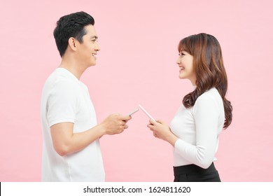 Face-to-face conversation. Excited man and woman talking holding smartphones looking at each other standing over pink Background.