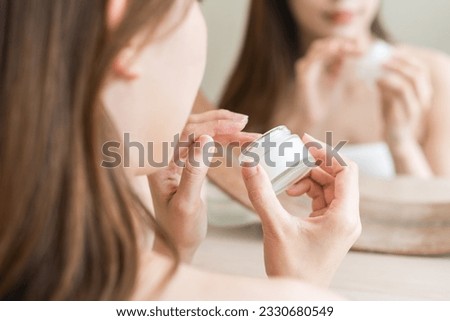 Facial beauty skin care, cute asian young woman, girl in bathrobe reflection into mirror, hand applying moisturizer lotion on her face, holding jar of skin cream before makeup cosmetic routine at home