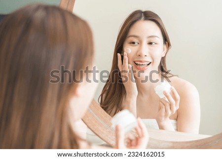 Facial beauty skin care, smile of pretty asian young woman in bathrobe looking at mirror, hand applying moisturizer lotion on her face, holding jar of skin cream before makeup cosmetic routine at home