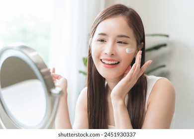 Facial beauty skin health care, smile of cute asian young woman looking at mirror, hand applying moisturizer lotion on her face, female putting cream treatment before makeup cosmetic routine at home. - Powered by Shutterstock