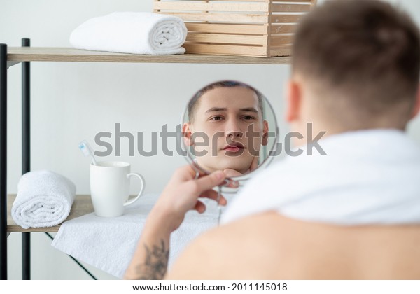 Facial care. Daily routine.\
Morning grooming. Aftershave lotion. Young handsome man touching\
smooth clean perfect face admiring mirror reflection in light\
bathroom.