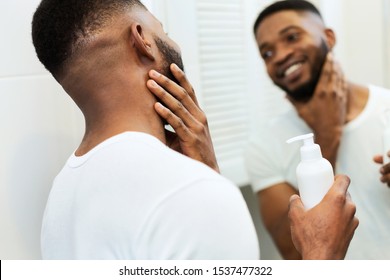 Facial Care. Young Black Man Applying Moisturizer On Beard In Bathroom, Looking In Mirror