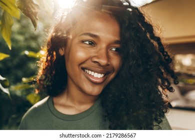 Facial close-up portrait of youthful African female walking down streets of favorite neighborhood, posing against green trees, smiling widely, feeling free, enjoying hot summer day