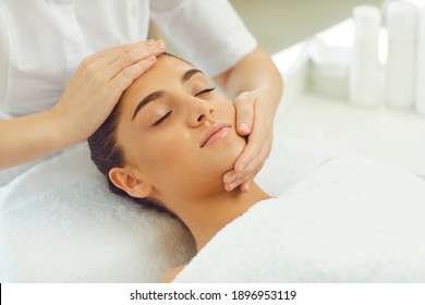 Facial massage in beauty wellness salon. Young positive relaxed woman getting procedure of facial massage from cosmetologists hands and enjoying fingers movements