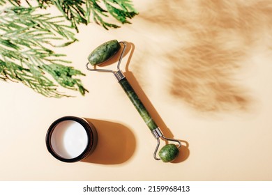 Facial Roller And Cream Jar With Cypress Branch Shadow On Light Beige Background. Natural Cosmetics, Aromatherapy, Beauty Treatment Concept. Top View, Flat Lay