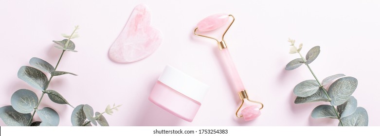 Facial roller, gua sha and moisturizing cream with green leaves, web banner. Facial tools on pink. Rose quartz beauty roller, gua sha flat lay. Tools for skin care routines, top view - Powered by Shutterstock