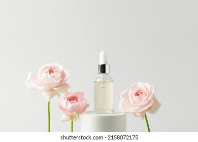 Facial serum in glass dropper bottle on white podium and pink roses on grey background. Natural skin care cosmetic. Beauty routine concept. Minimal composition. Trendy product for branding.