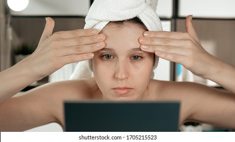 Facial skin massage. Attractive girl in bathroom with towel and mirror does facial massage on her forehead with her hands. Hygiene, beautician, makeup, face care concept. Close-up
