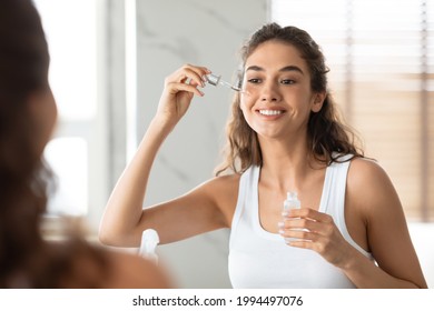 Facial Skincare. Attractive Female Applying Serum On Face Moisturizing And Caring For Skin Standing Near Mirror In Modern Bathroom Indoors. Beauty Routine. Skin Care Concept. Selective Focus