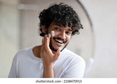 Facial skincare. Indian man applying moisturizing cream on face, looking at his reflection in bathroom mirror at home, free space. Male beauty and skin care concept - Powered by Shutterstock