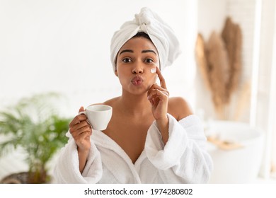 Facial Skincare. Playful Black Woman Applying Cream On Face Holding Cup Of Coffee Posing Pouting Lips In Modern Bathroom At Home. Morning Beauty Routine Concept