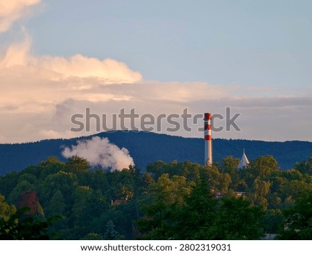 Facility chimney rises beside dense trees on wooded hillside while pink and blue evening clouds glow overhead light smoke drifts into calm air, contrasting heavy industry with natural scenery