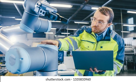 At The Factory: Automation Engineer Uses Laptop For Programming Robotic Arm. New Era In Automatic Manufacturing Industry.