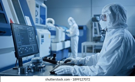 Factory Cleanroom: Engineer Wearing Coverall And Mask Does System Control On A Computer, Uses Controller, Screen Shows Infographics. In Background Modern CNC Machinery, Robot Arm, Electronic Equipment