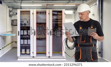 Factory electrician. Man from energy company. Electrification of production. Electrician guy with tablet and documentation. Electrical cabinet is behind man. Electrician in technical room of factory