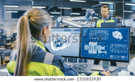 At the Factory: Female Mechanical Engineer Designs 3D Engine on Her Personal Computer. In the Background Male Automation Engineer who Uses Laptop for Programming Robotic Arm.