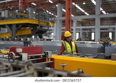 In a factory setting, workers are managing large machines. They wear safety helmets and vests. The area is bright and busy with various equipment.
