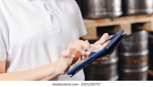 Factory worker with computer tablet inspecting kegs with beer in warehouse of modern brewery stock, blurred background.